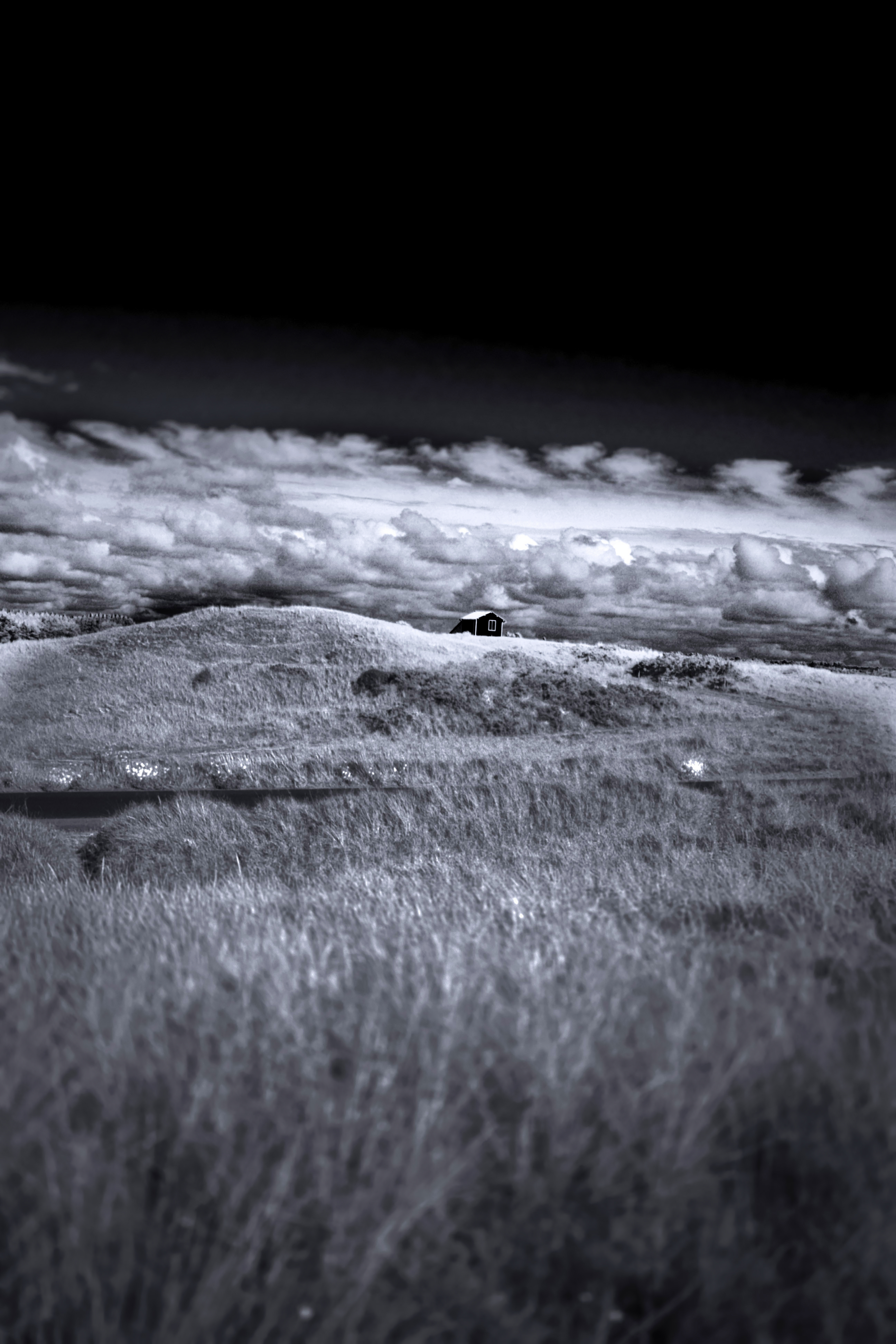 Infrared landscape with a lone cabin on grassy hills under a dramatic black sky